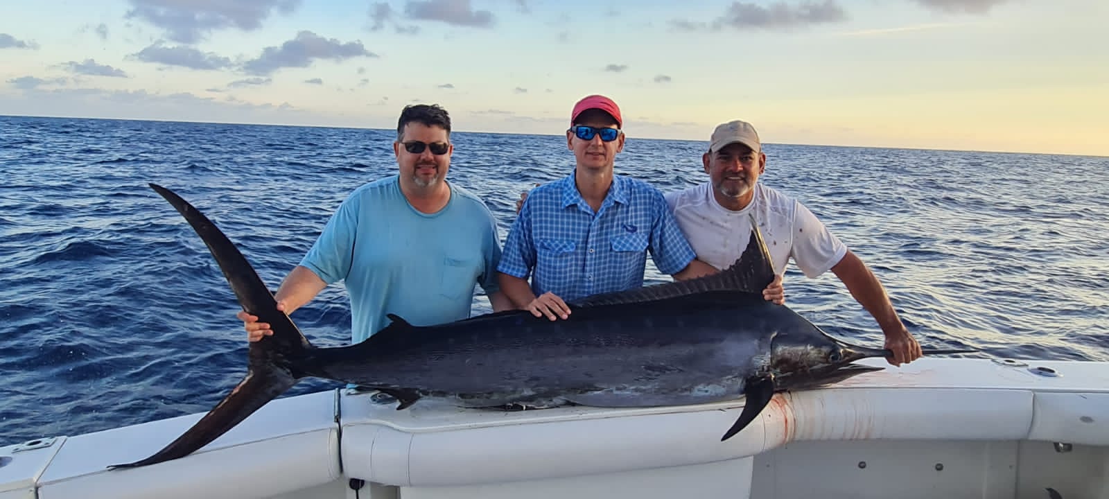 Three men on a fishing boat at sea holding a large blue marlin across the stern