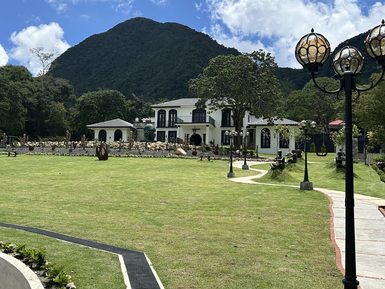 A white colonial-style house with a lawn and lamp posts below a green mountain